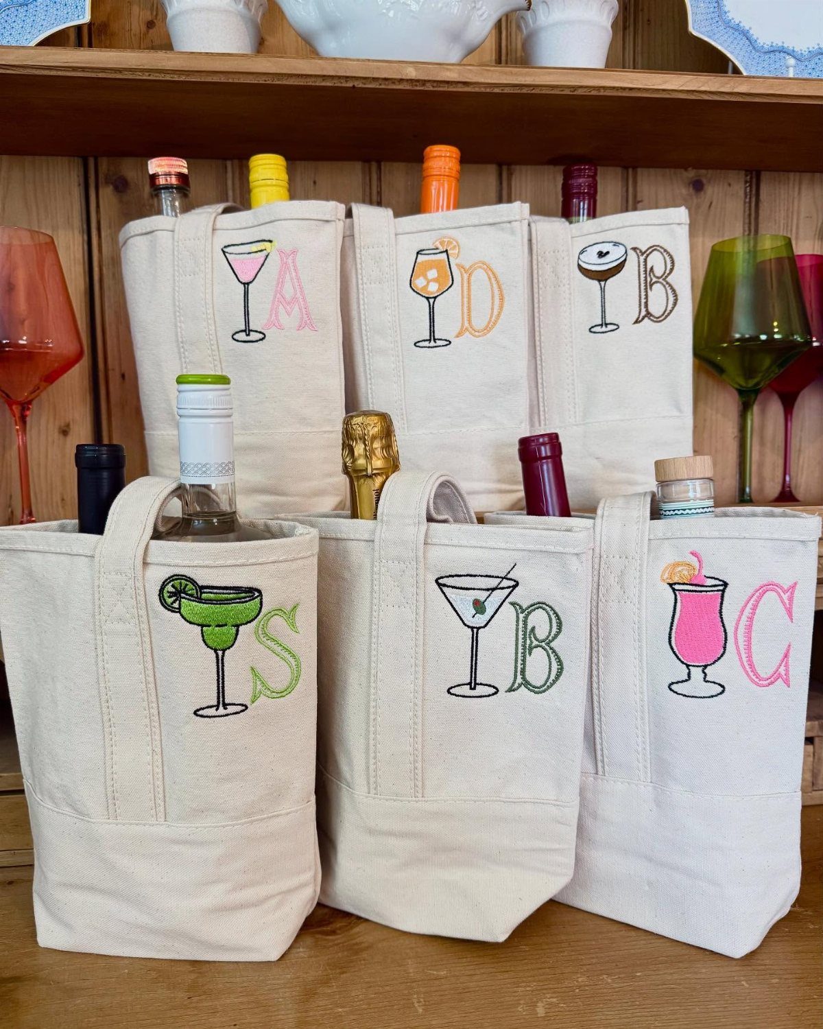 Canvas wine tote bags with embroidered cocktail glasses and personalized initials on wooden shelf