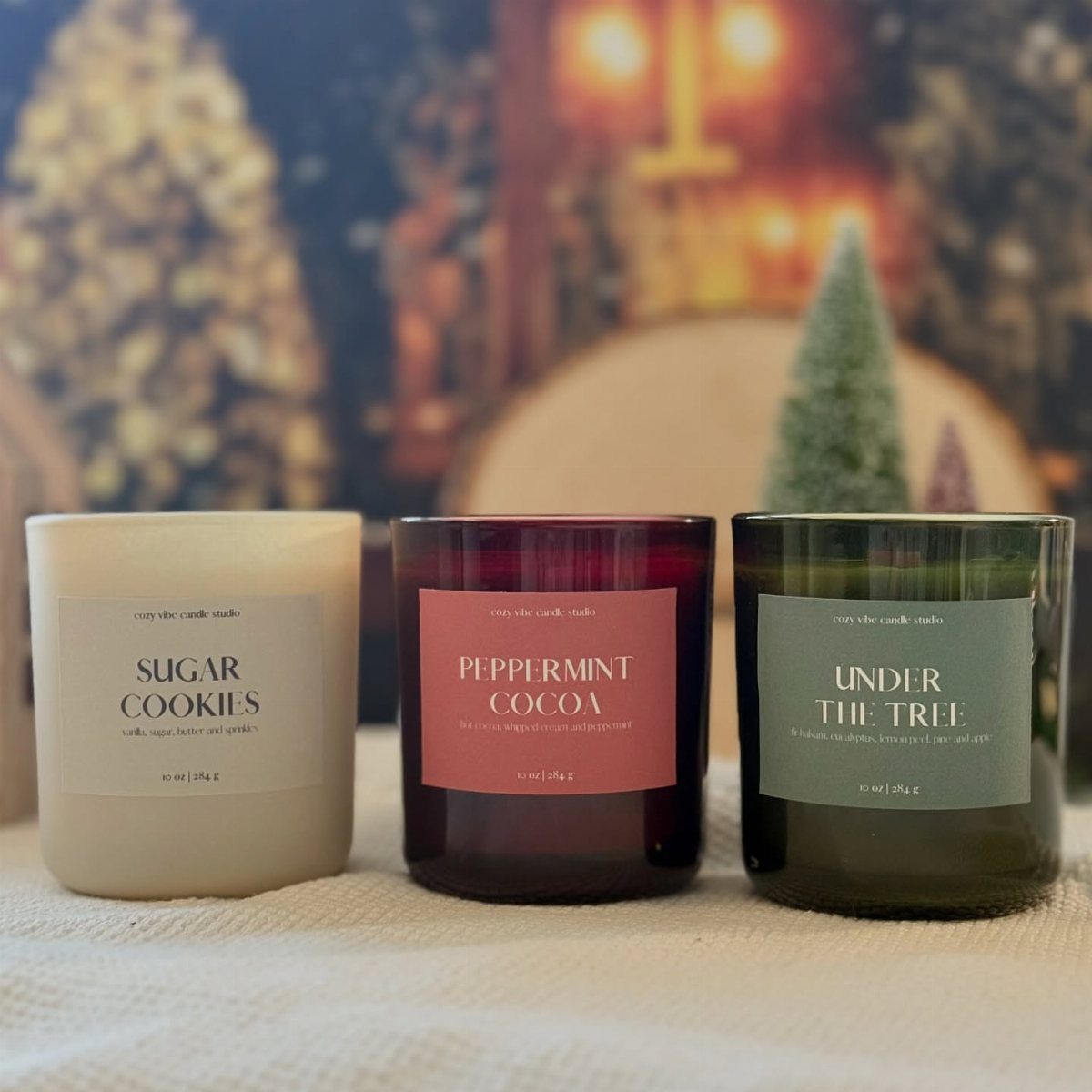 Three holiday scented candles in glass jars on a white fabric surface.