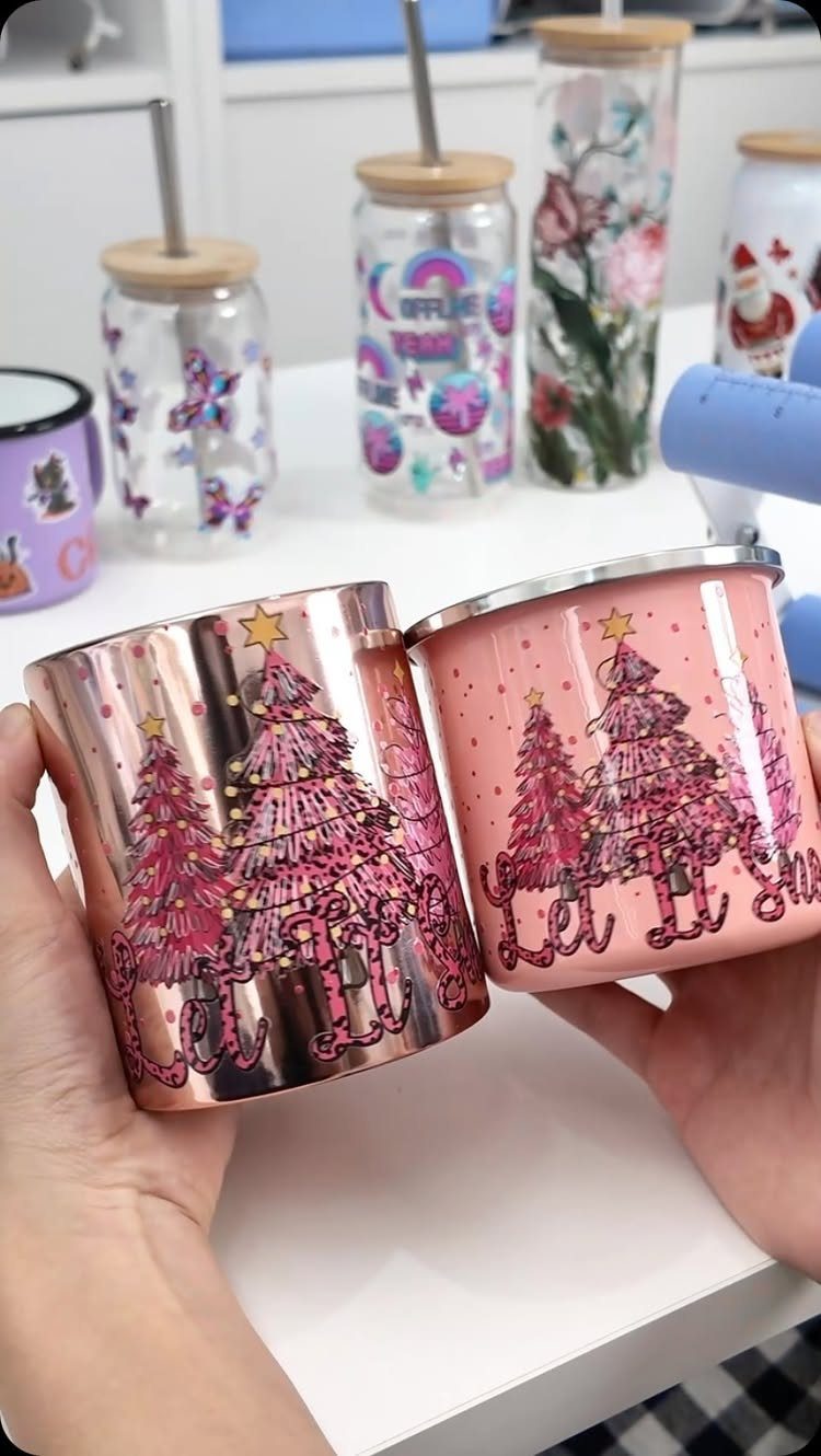 Two metallic tumblers with pink Christmas tree designs held by two hands.