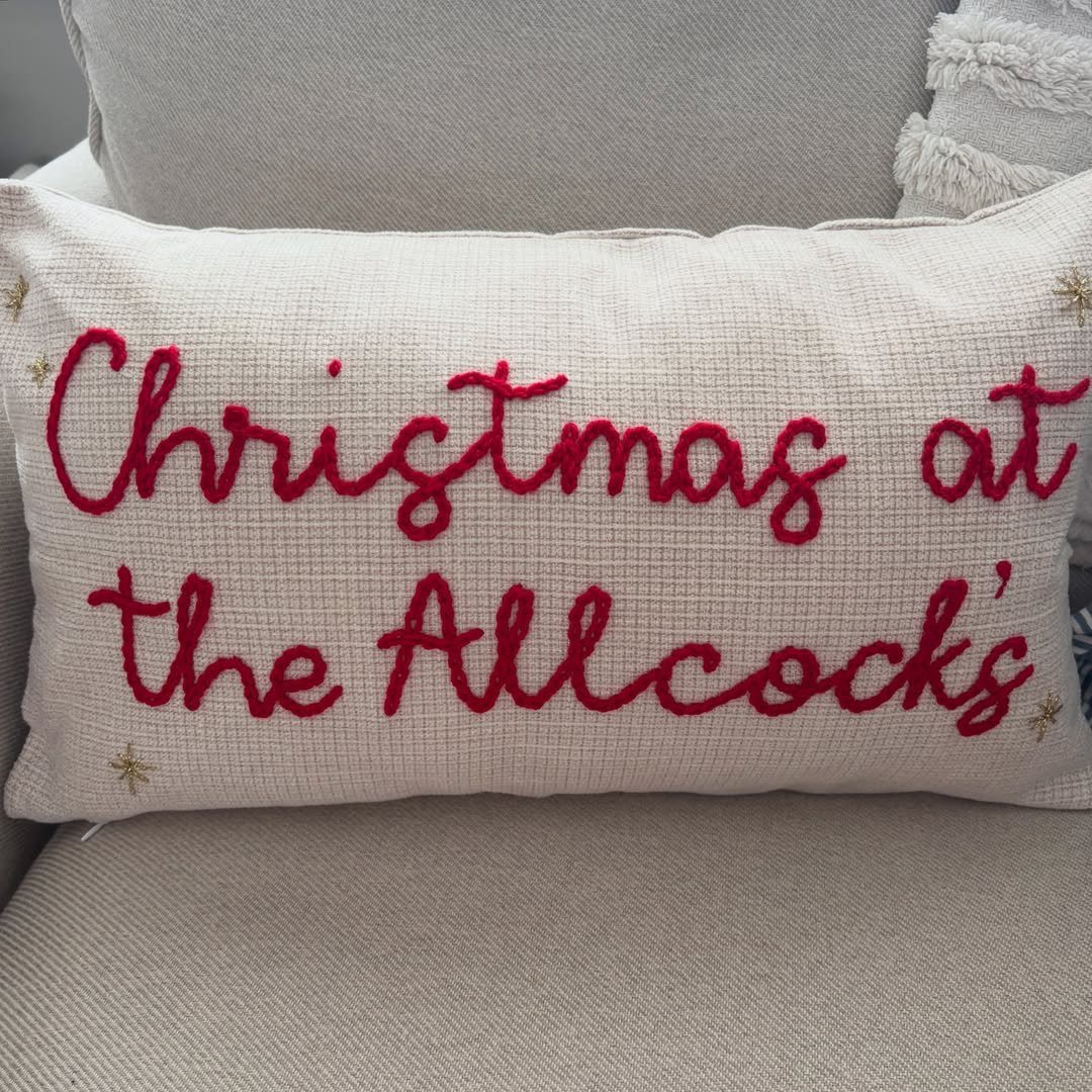 Cream throw pillow with red embroidered text Christmas at the Allcock's on sofa