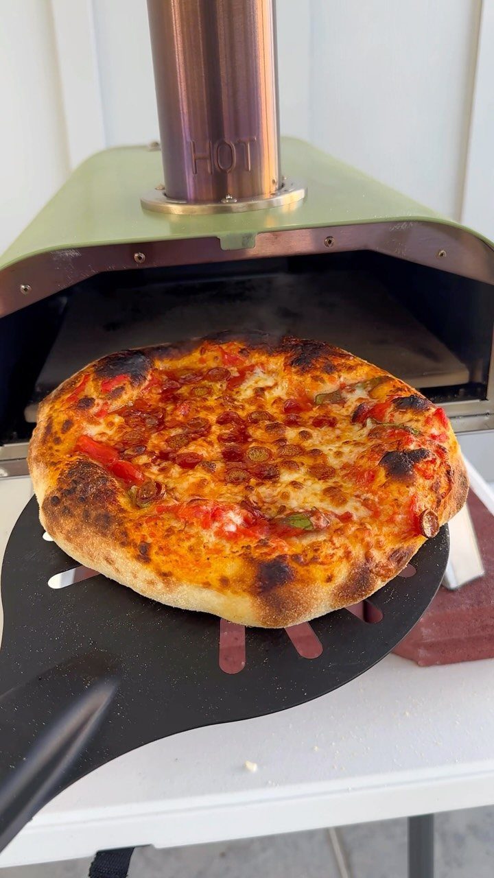 Freshly baked pizza on a peel being removed from a green outdoor oven.