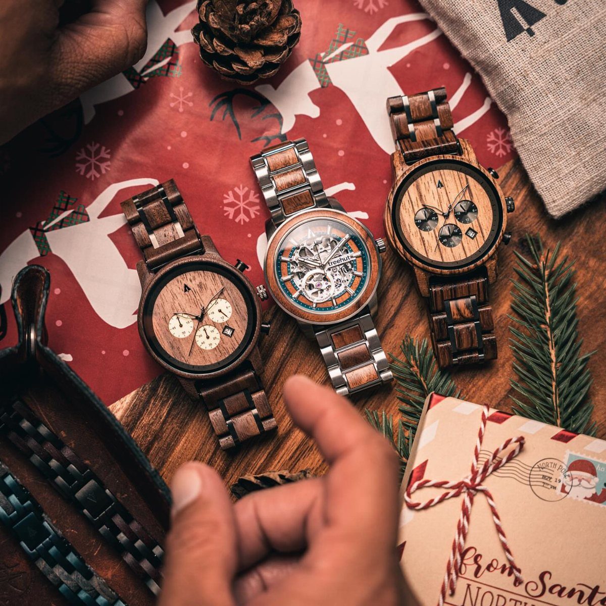 Three wooden wristwatches on red Christmas wrapping paper with pine cones and gifts.