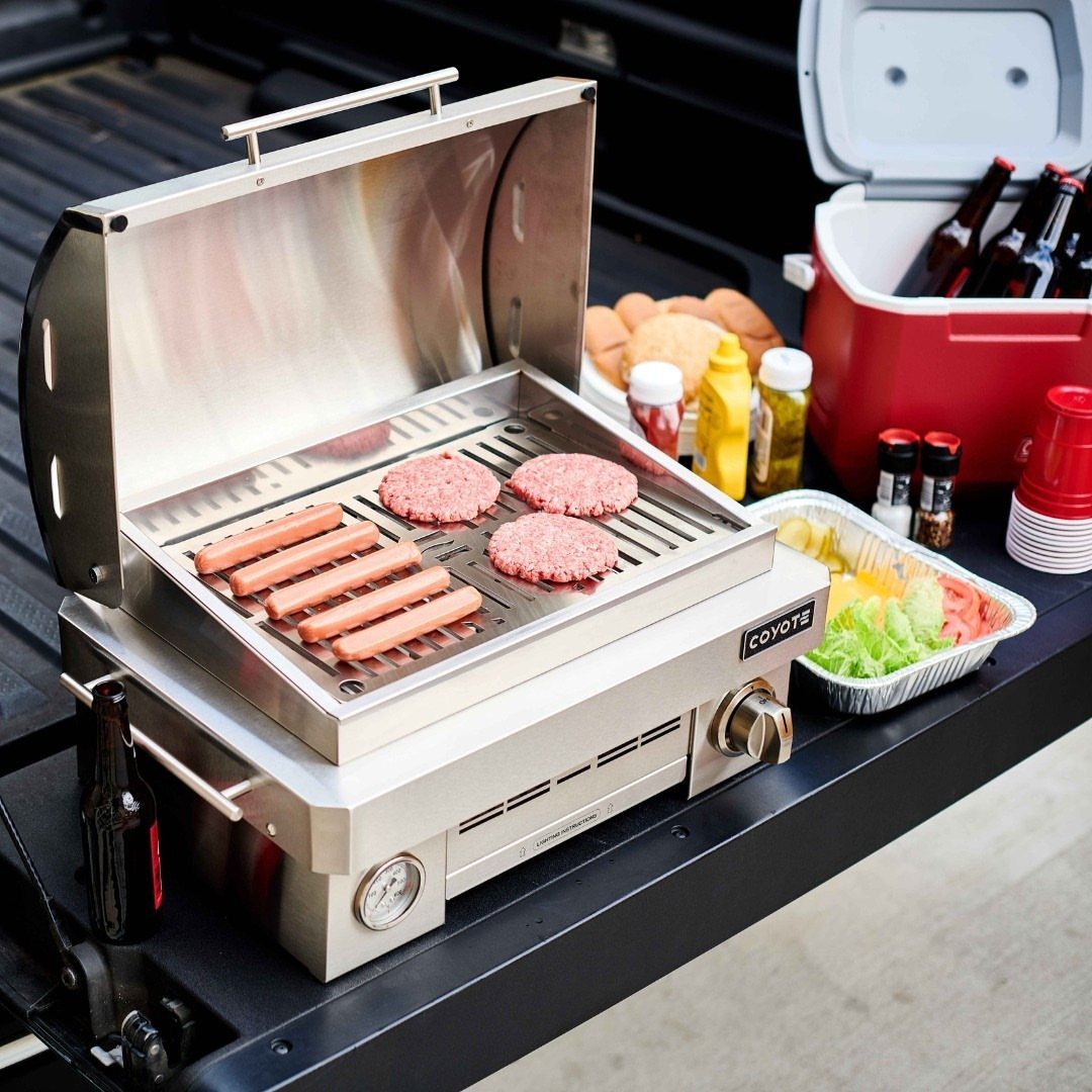 Portable stainless steel grill cooking burgers and sausages on a truck tailgate.