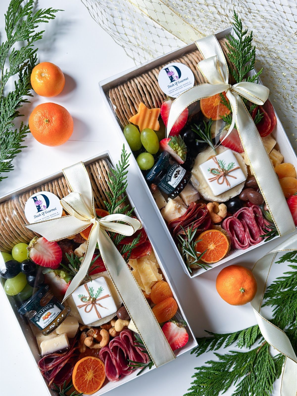 Two charcuterie gift boxes with ribbons, tangerines, and cedar branches on white background