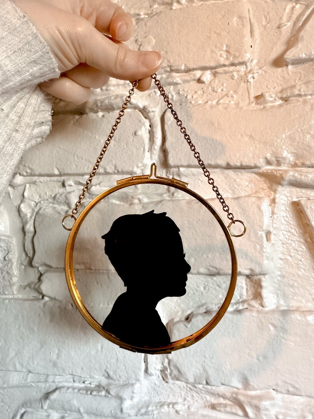 Gold circular frame with a black silhouette profile held against white brick wall