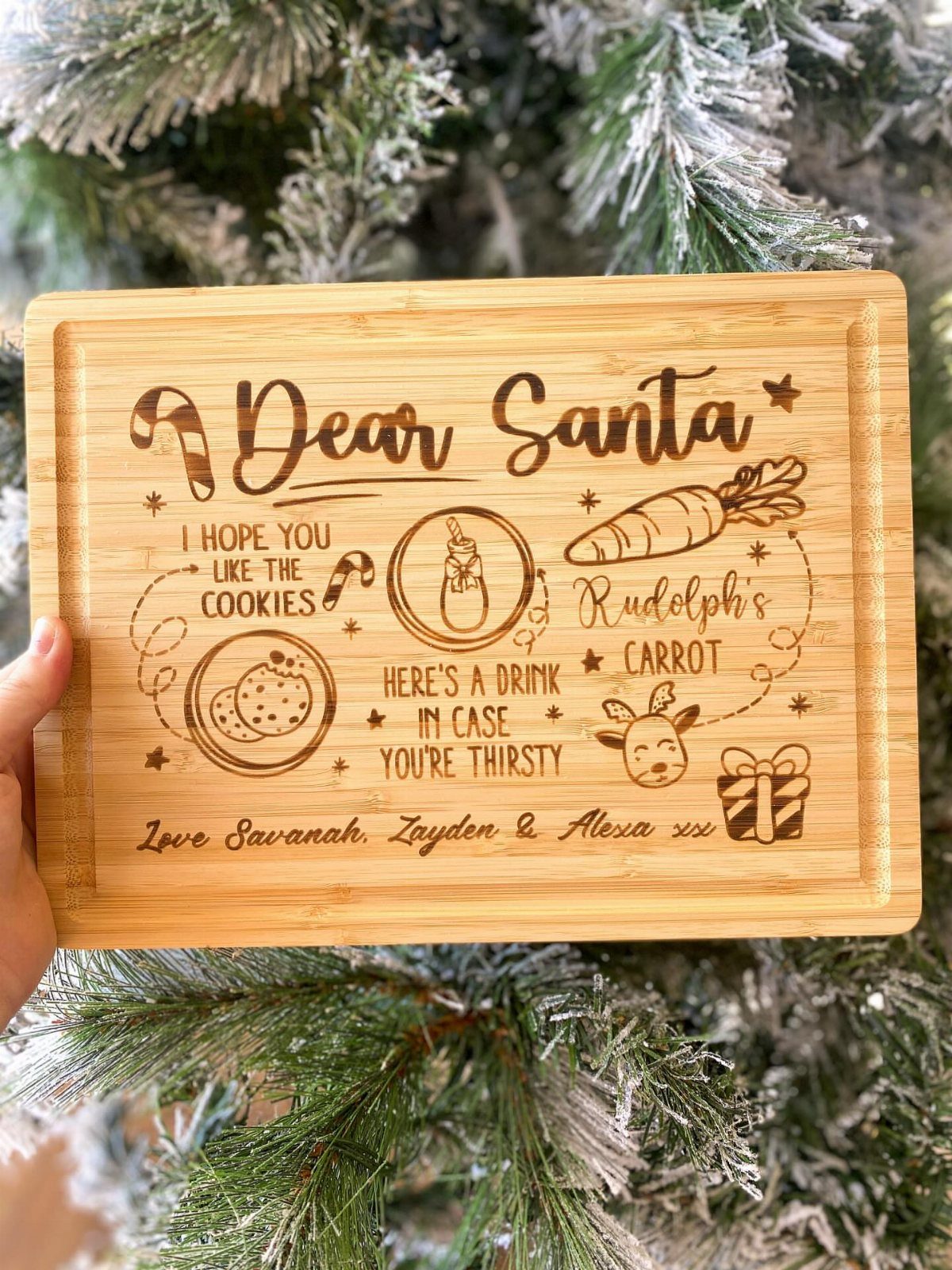 Engraved bamboo cutting board for Santa with cookies and carrots against frosted pine.