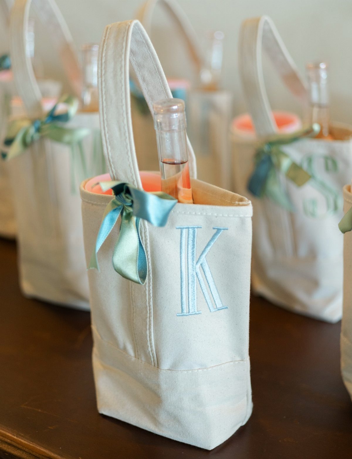 Cream canvas wine tote bags with blue bows and embroidered letters holding rosé bottles.