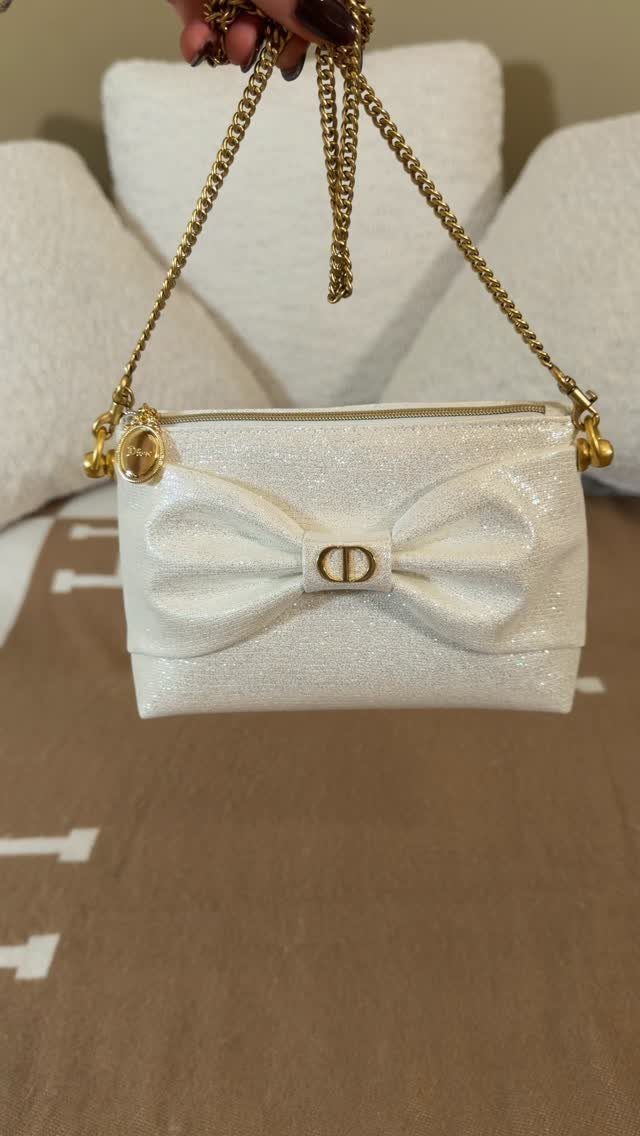 White shimmering handbag with a large bow and gold chain strap held by hand.