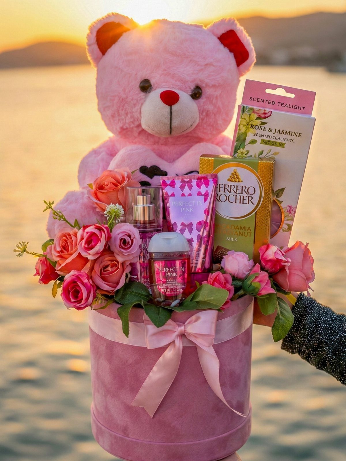 Pink gift box with teddy bear, roses, chocolates, and beauty products at sunset.