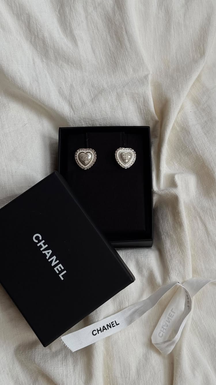 Heart-shaped silver earrings in a black Chanel jewelry box on white fabric.