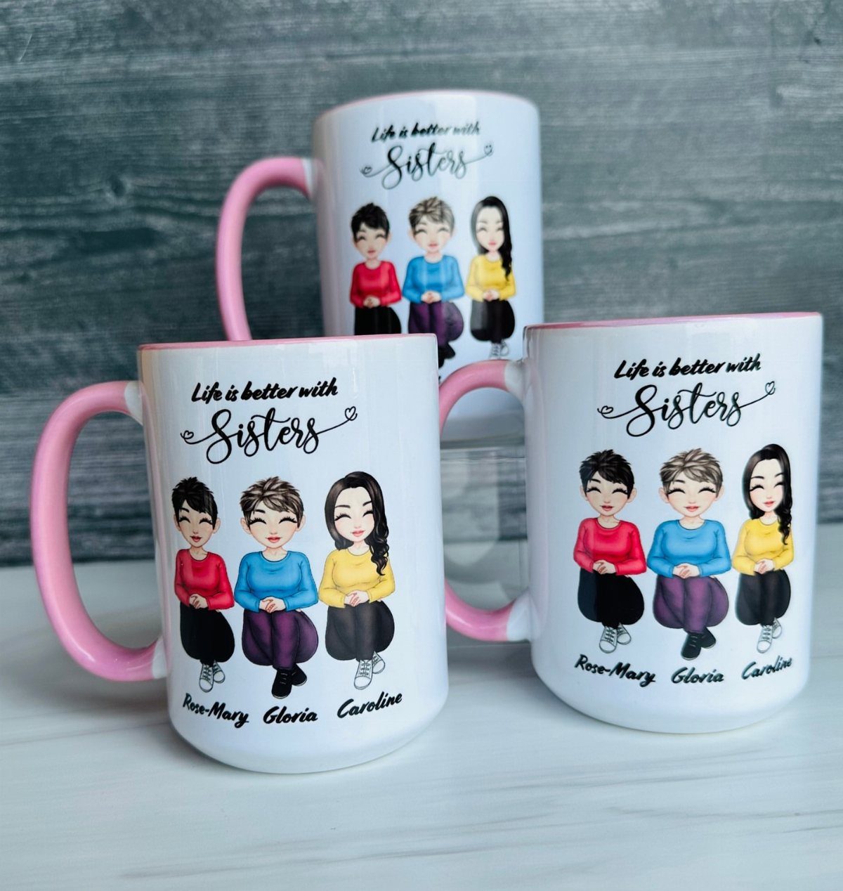 Three personalized white mugs with pink handles featuring sister illustrations and names.