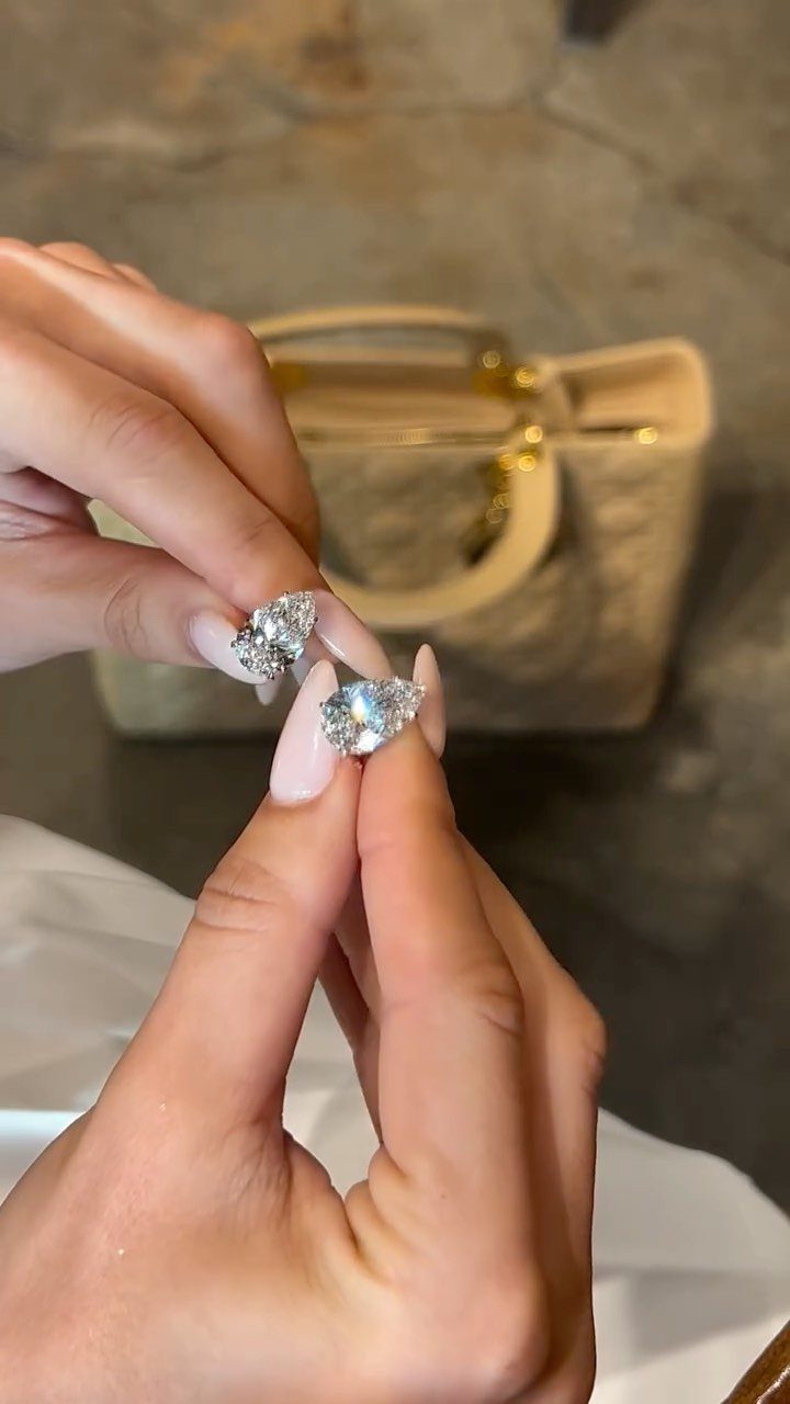 Hands holding two sparkling diamond earrings with a beige handbag in background