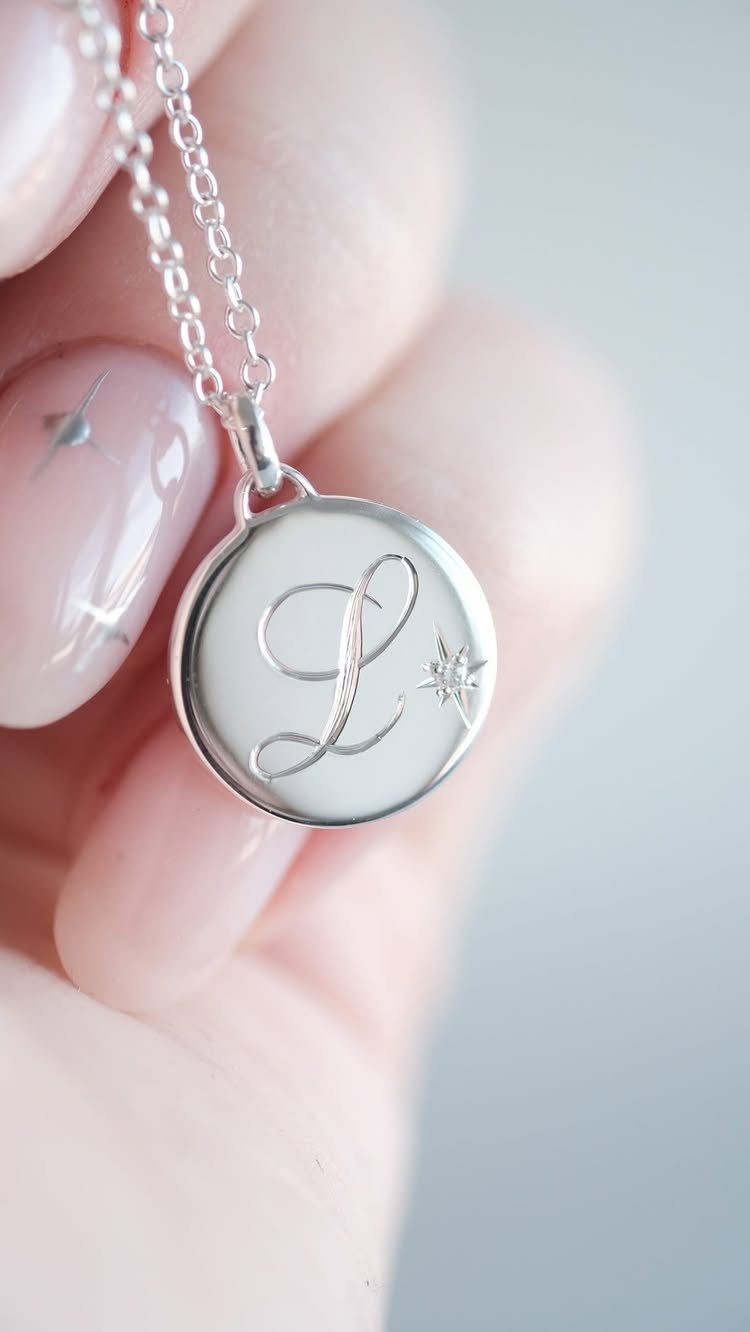 Silver circular pendant necklace engraved with a cursive letter L held by hand