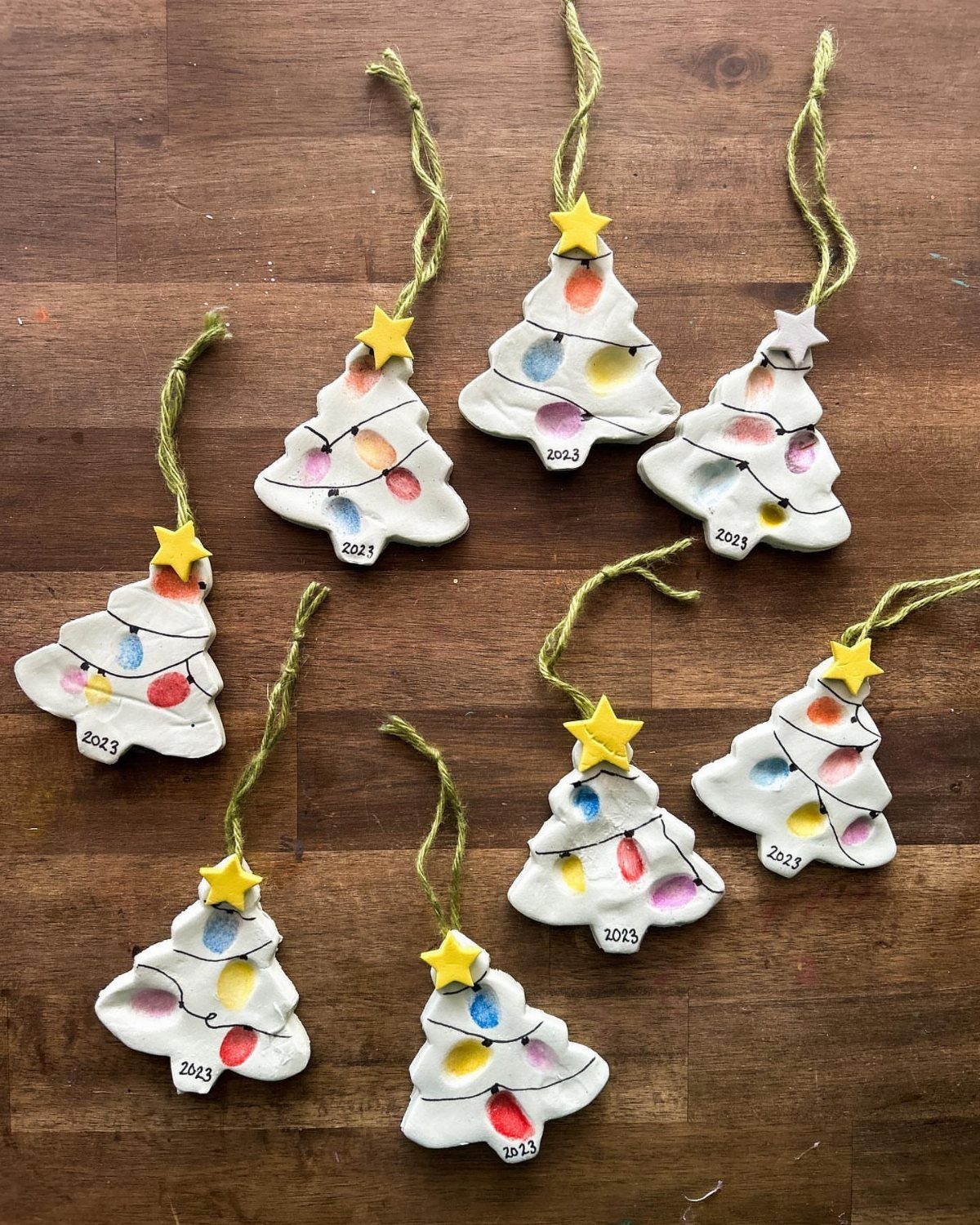 White clay Christmas tree ornaments with colorful lights and yellow stars on wood.