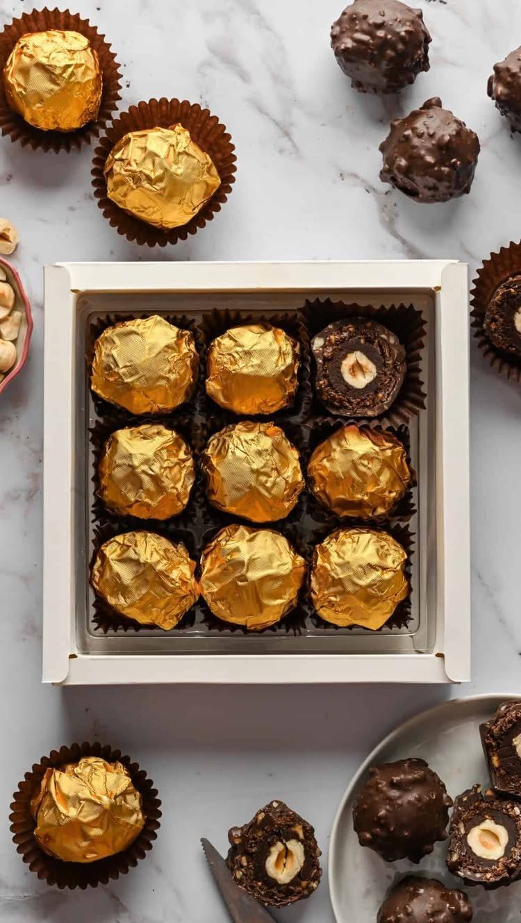 Box of gold-wrapped chocolate truffles and dark chocolate hazelnut balls on marble surface