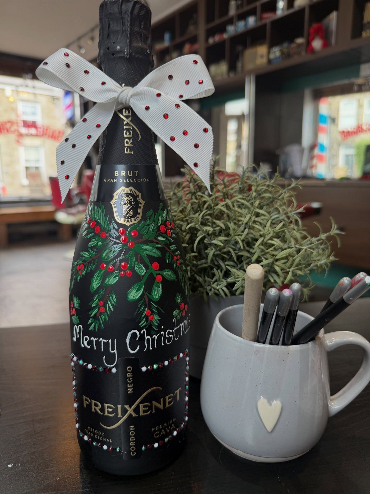 Decorated champagne bottle with painted holly and a white bow next to brushes.