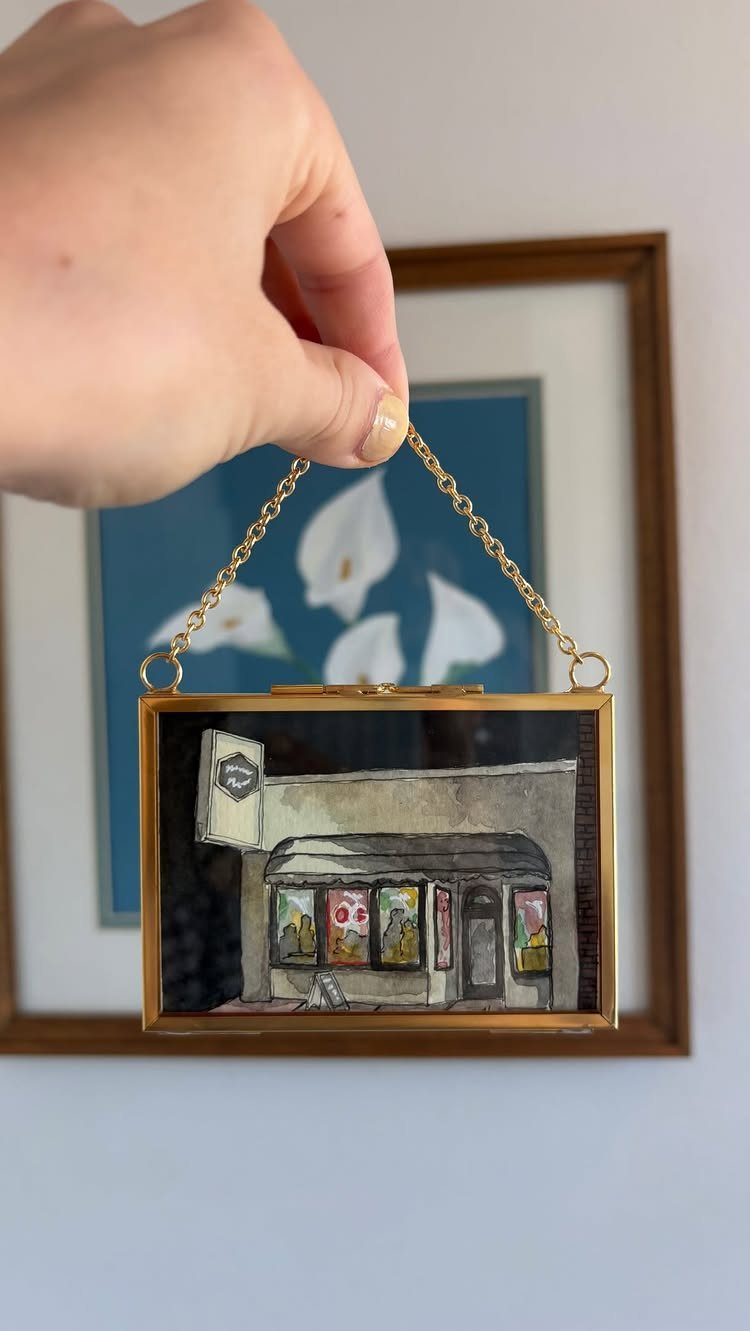 Small gold-framed painting of a storefront held by a gold chain.
