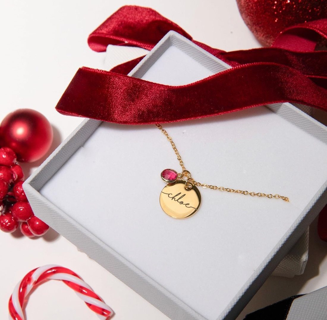 Gold personalized name necklace in a white box with Christmas decorations around it.