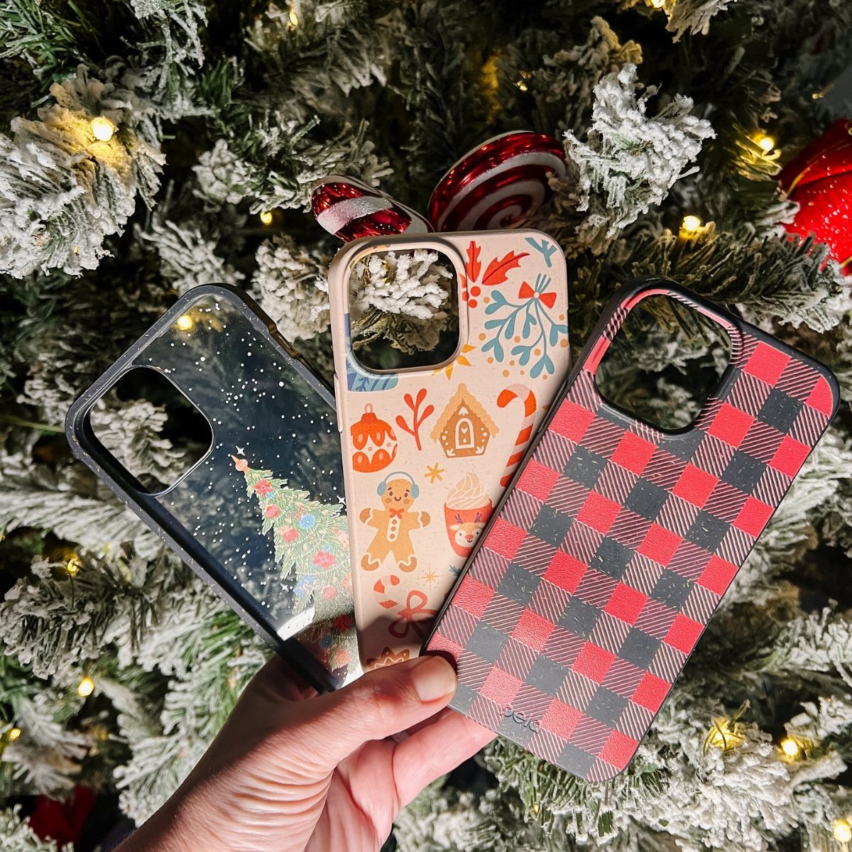 Three festive phone cases held against a lit, snowy Christmas tree background.