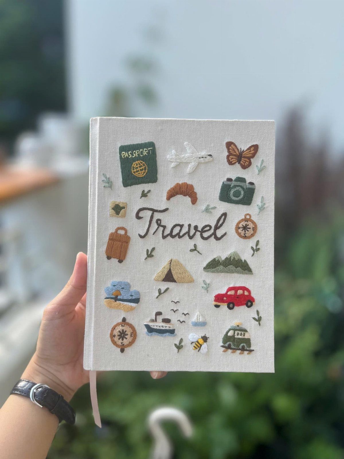 White fabric travel journal with embroidered travel icons held by a person's hand.