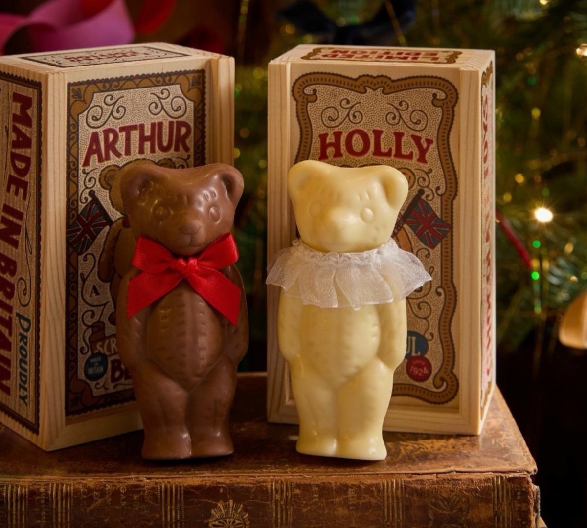 Two chocolate bears with bows standing before their decorative gift boxes on a book.