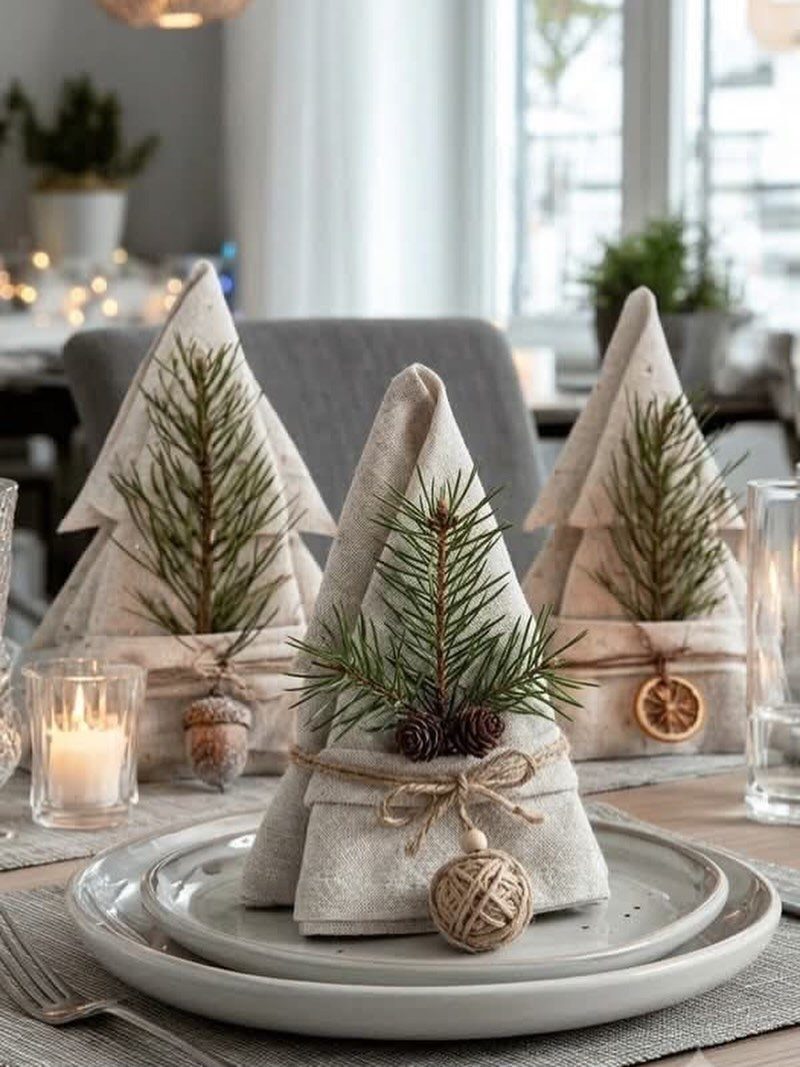 White plates with beige cloth napkins folded as trees and pine branch accents.