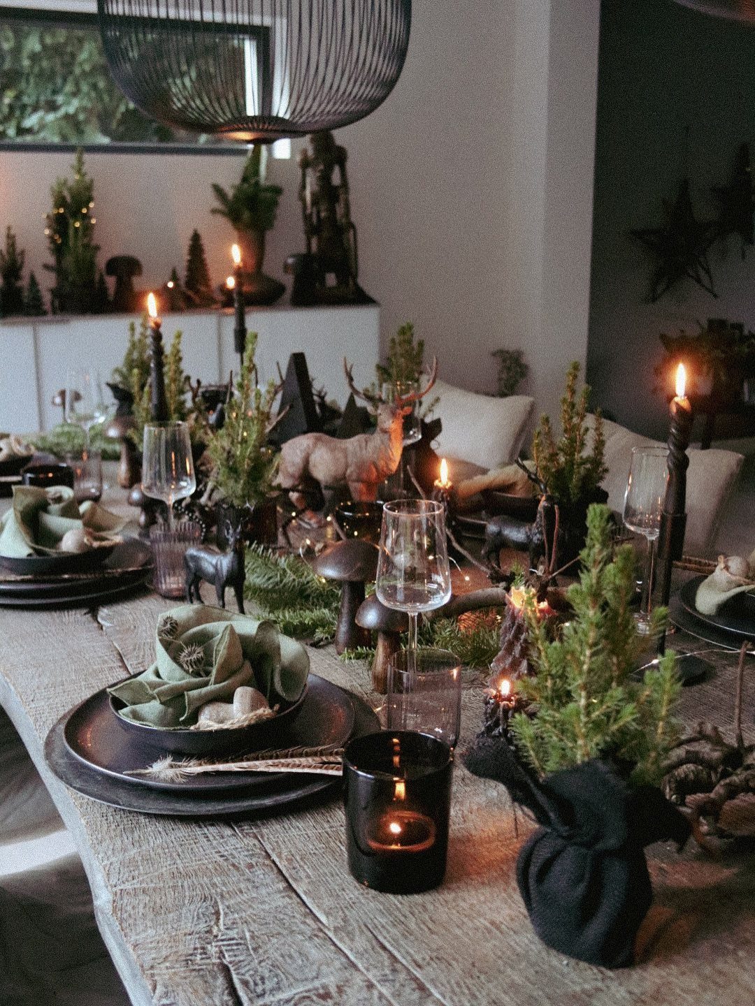Moody Christmas tablescape with black plates, evergreen trees, and candles on rustic wood.