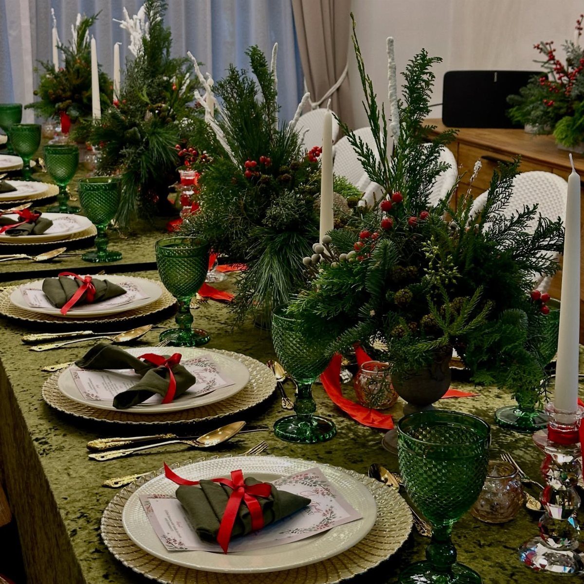 Festive dining table with green glassware, evergreen centerpieces, and red ribbon napkins.