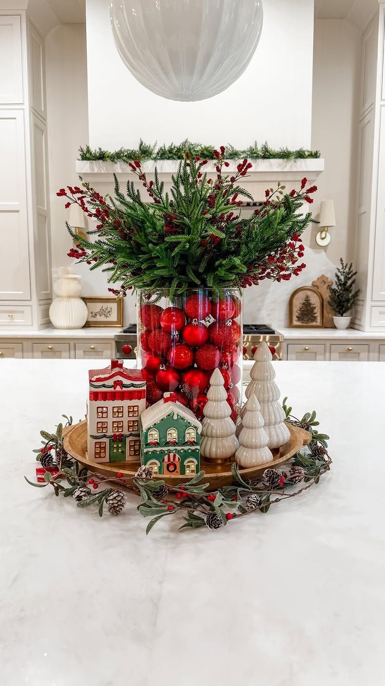 Christmas centerpiece with red ornaments in a vase, greenery, and miniature houses.
