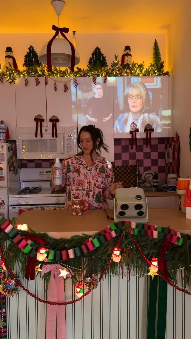 Woman in festive pajamas in a kitchen filled with dense Christmas greenery and decorations