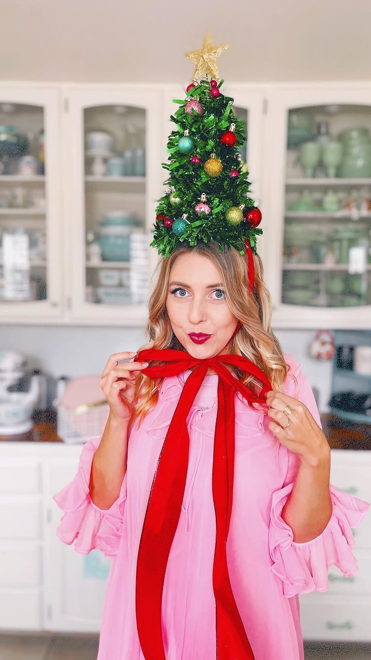 Woman wearing a green Christmas tree hat with ornaments and a red ribbon.
