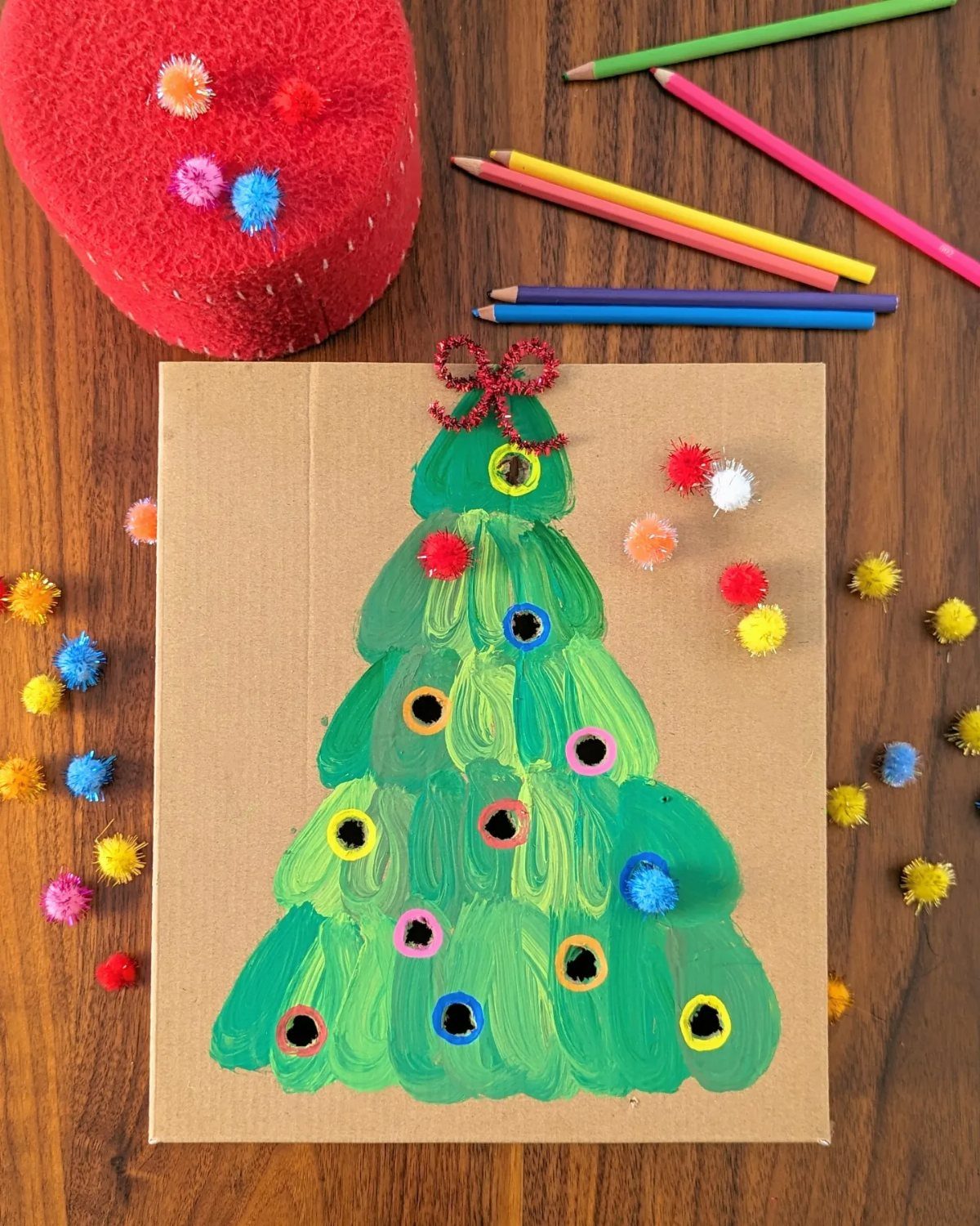 Green painted Christmas tree on brown card decorated with colorful pom poms.