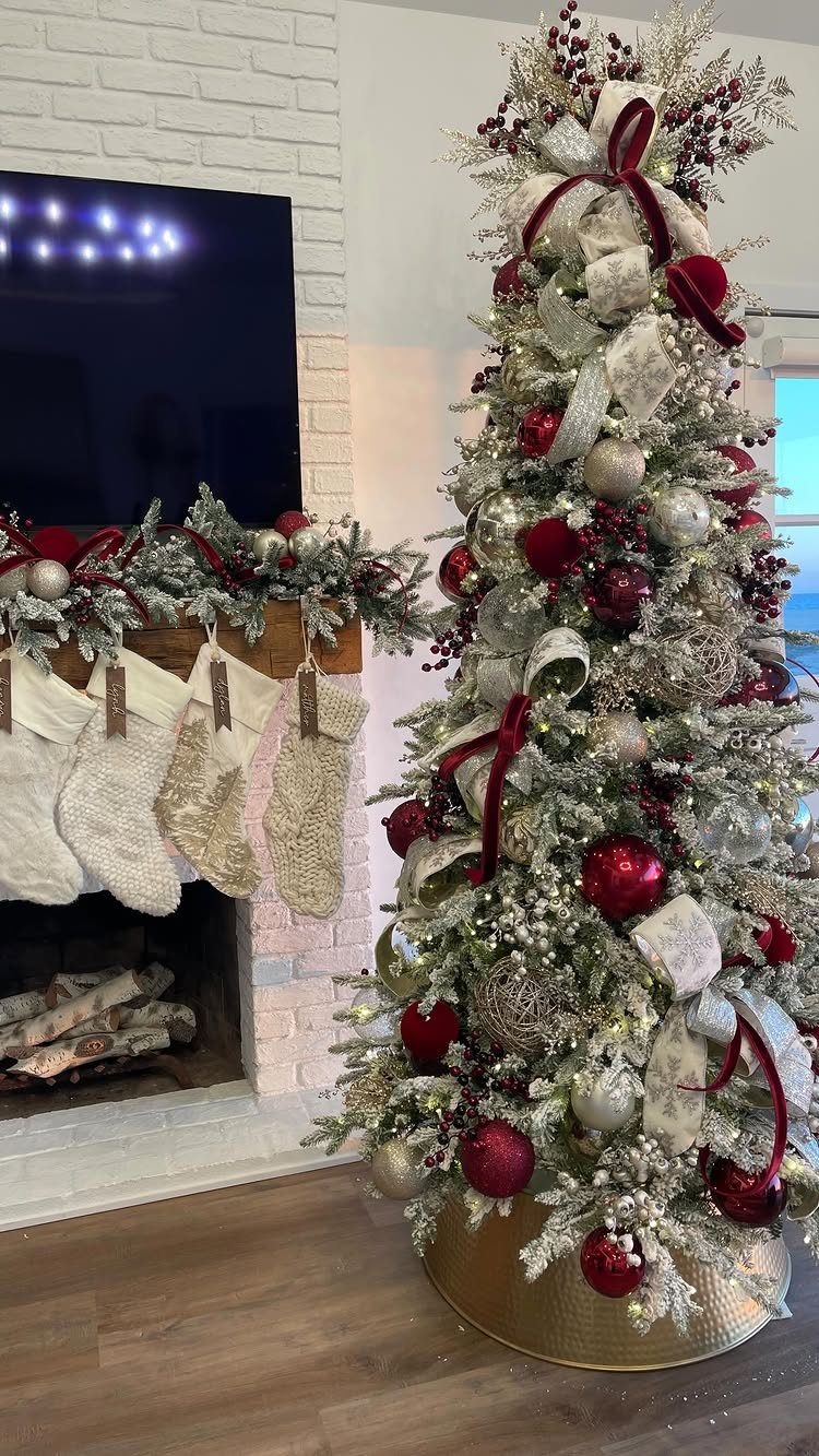 Christmas tree decorated with red and silver ornaments and matching ribbons by a fireplace.
