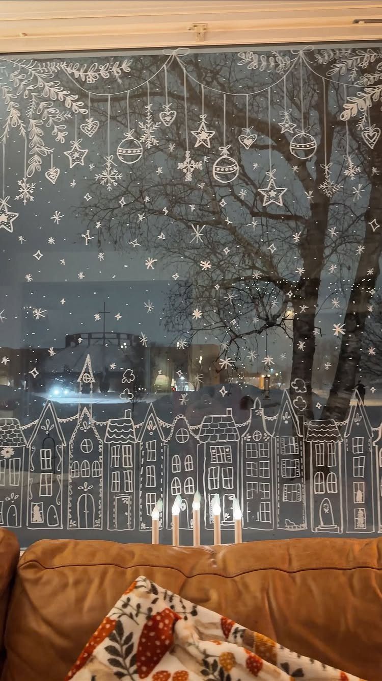 Window decorated with white drawings of a Christmas village and hanging ornaments.