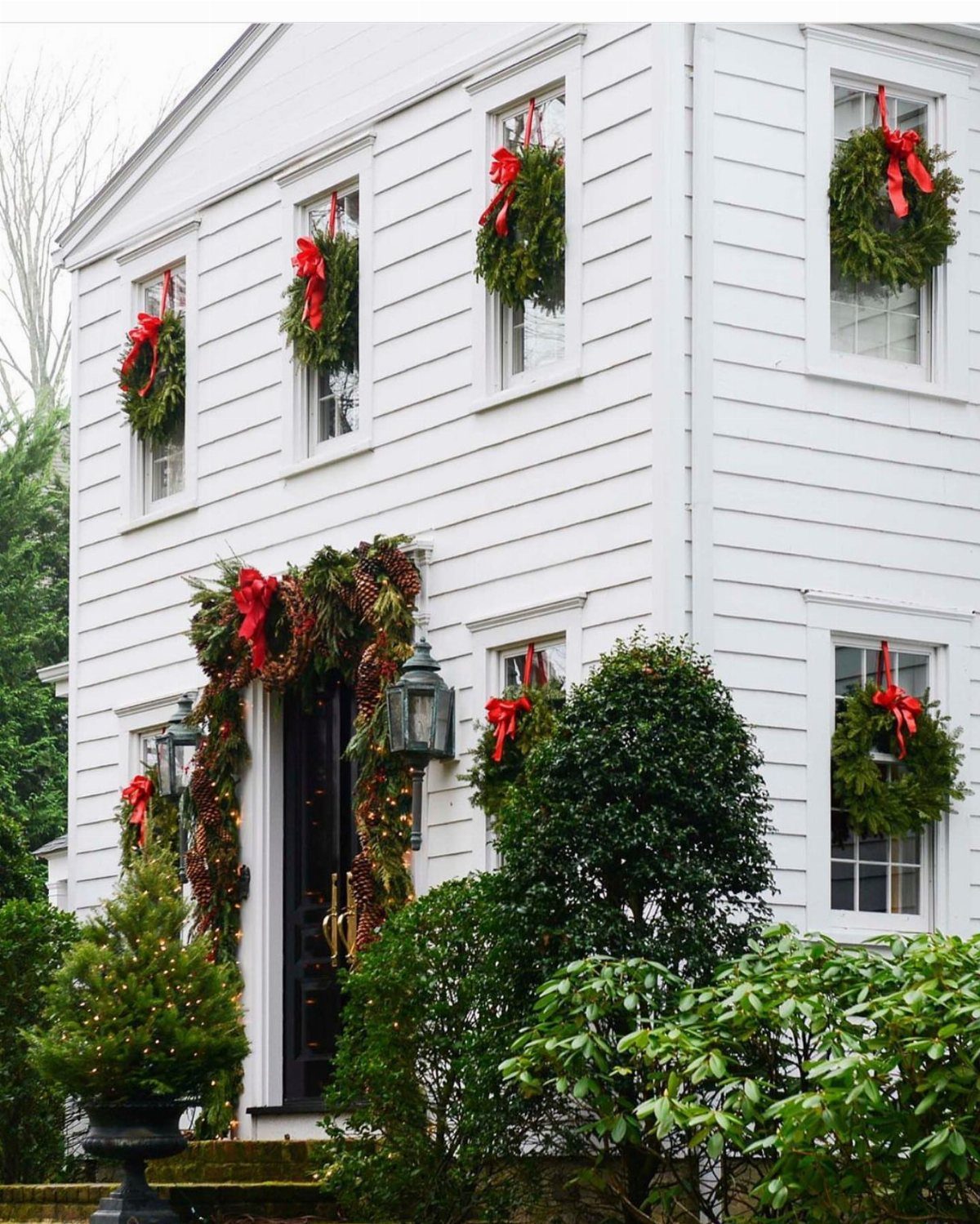 Green Christmas wreaths with red bows hanging in the windows of a white house.