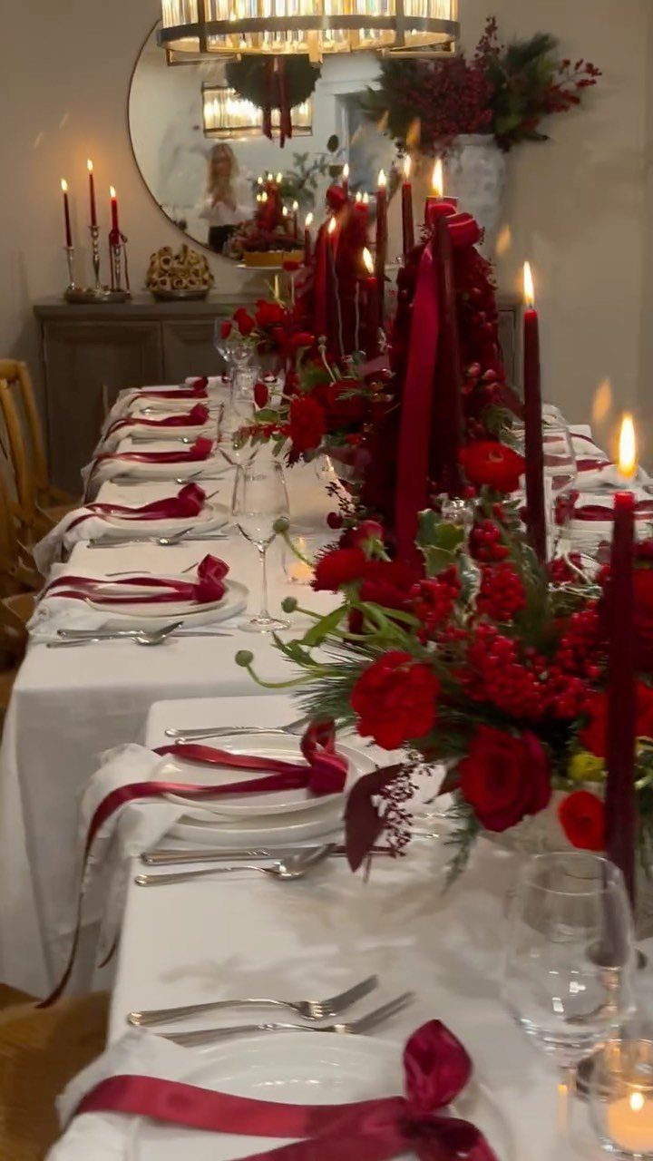 Dining table centerpiece with red flowers, green foliage, and tall red taper candles.