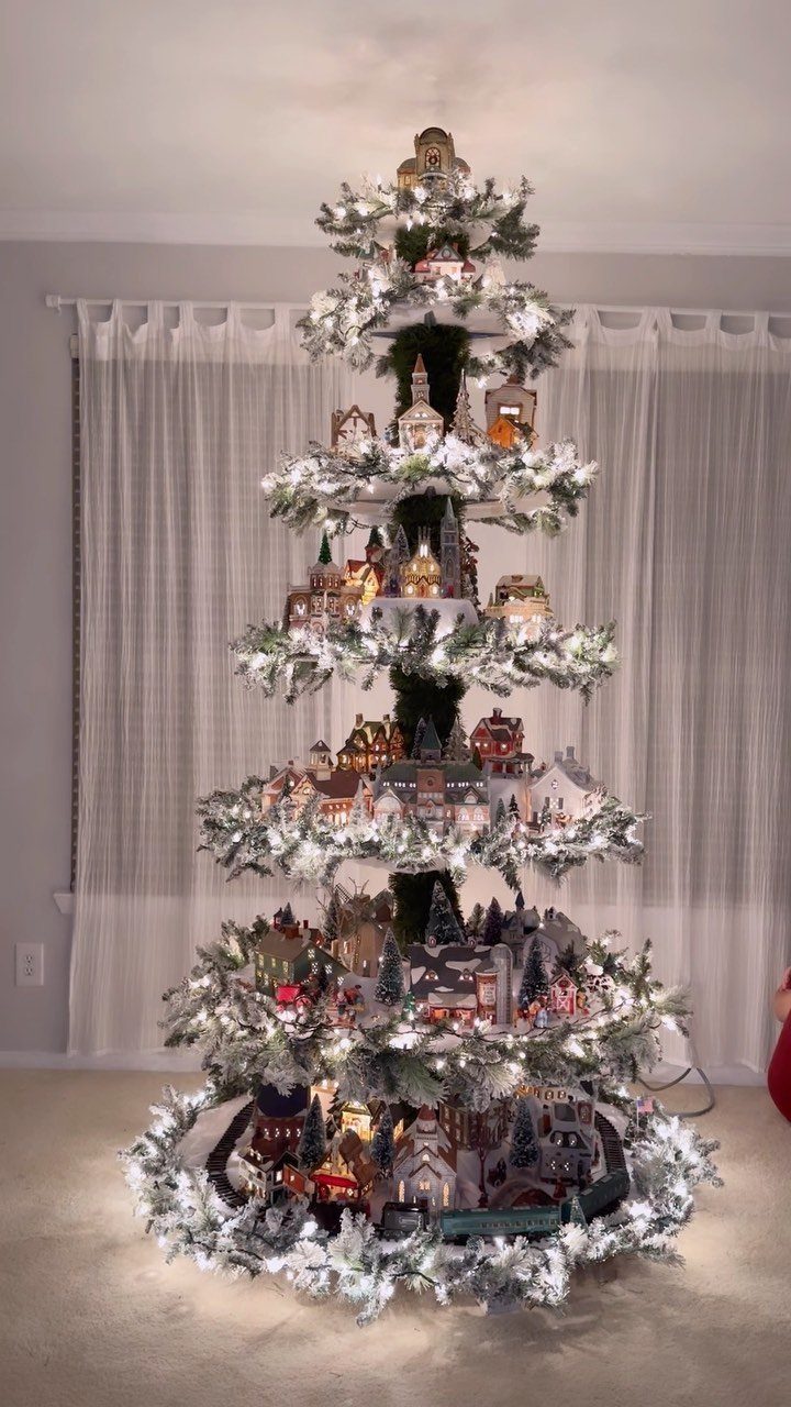 Tiered snow-covered Christmas tree featuring miniature illuminated village houses on every level.