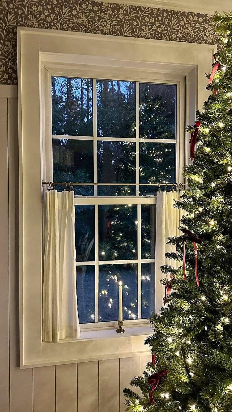 Lit white candle on a windowsill next to a decorated Christmas tree at dusk