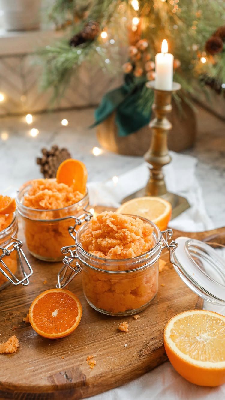 Orange body scrub in glass jars on a wooden board with citrus slices.