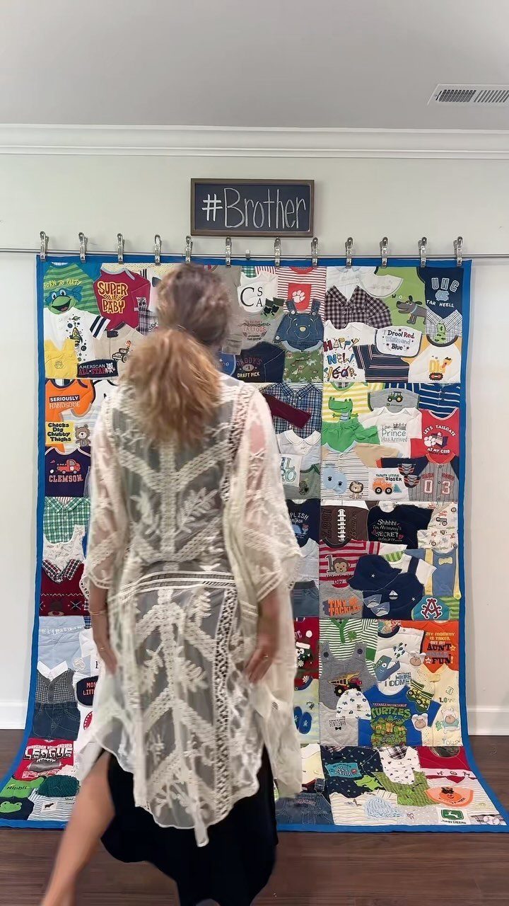 Woman looking at a colorful memory quilt made from baby clothes hanging on wall.