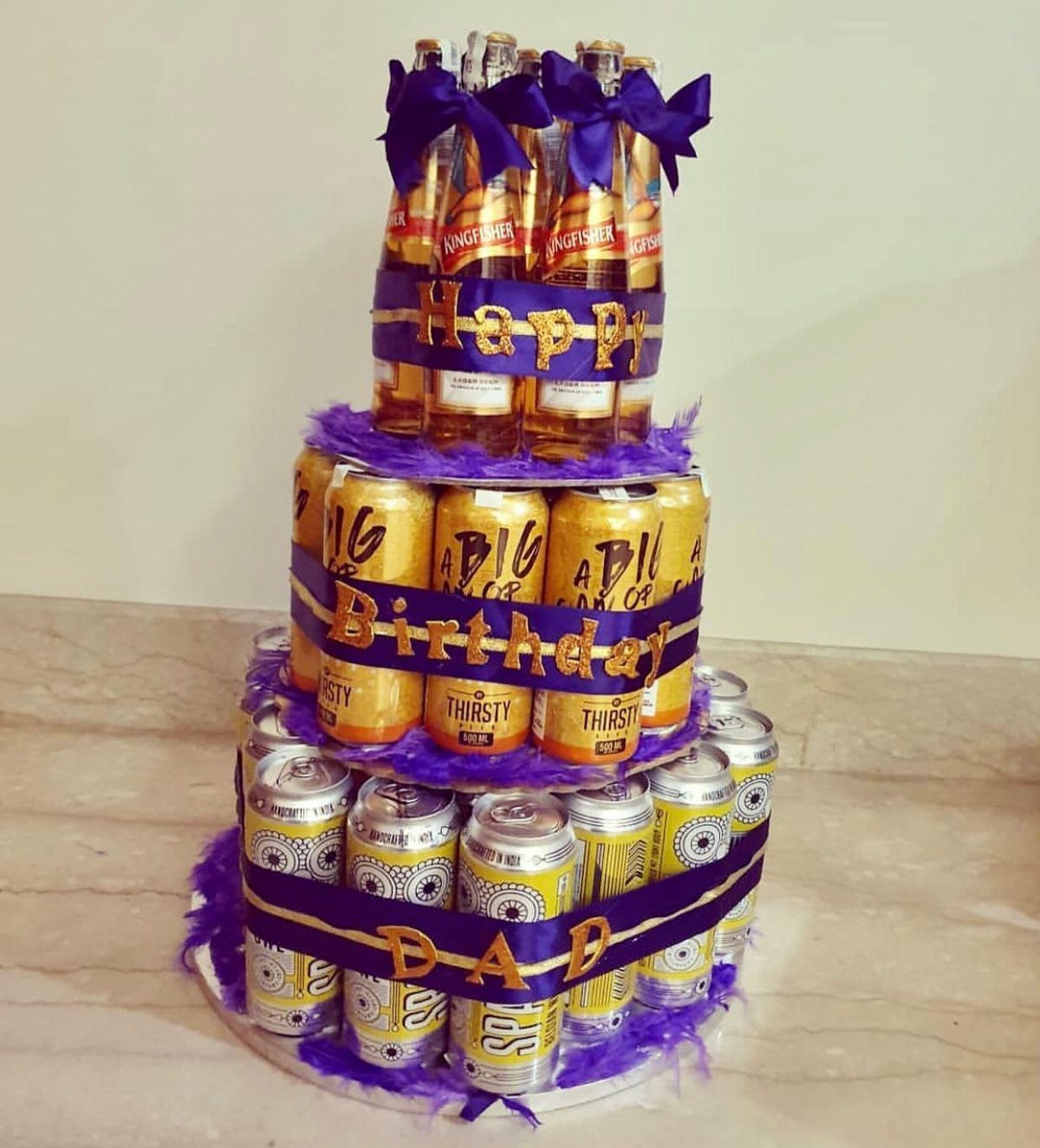 Beer bottle and can cake decorated with purple ribbons and gold birthday text.