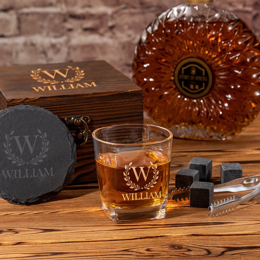 Engraved whiskey glass with amber liquid, wooden box, and crystal decanter on wood.