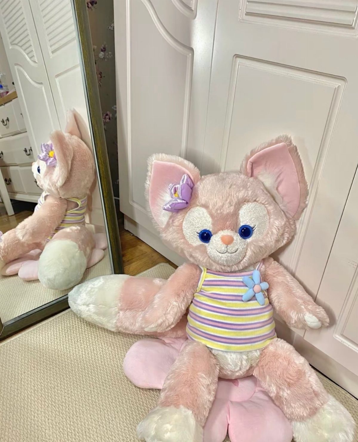 Pink plush cat toy in a striped shirt sitting beside a mirror.