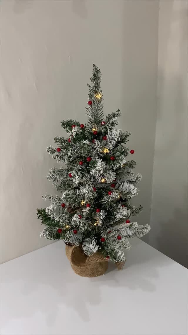 Small snow-dusted Christmas tree with red berries and lights on a white table