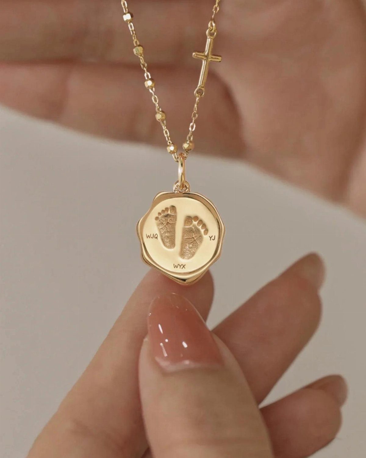 Gold necklace with a pendant engraved with baby footprints and initials held by hand