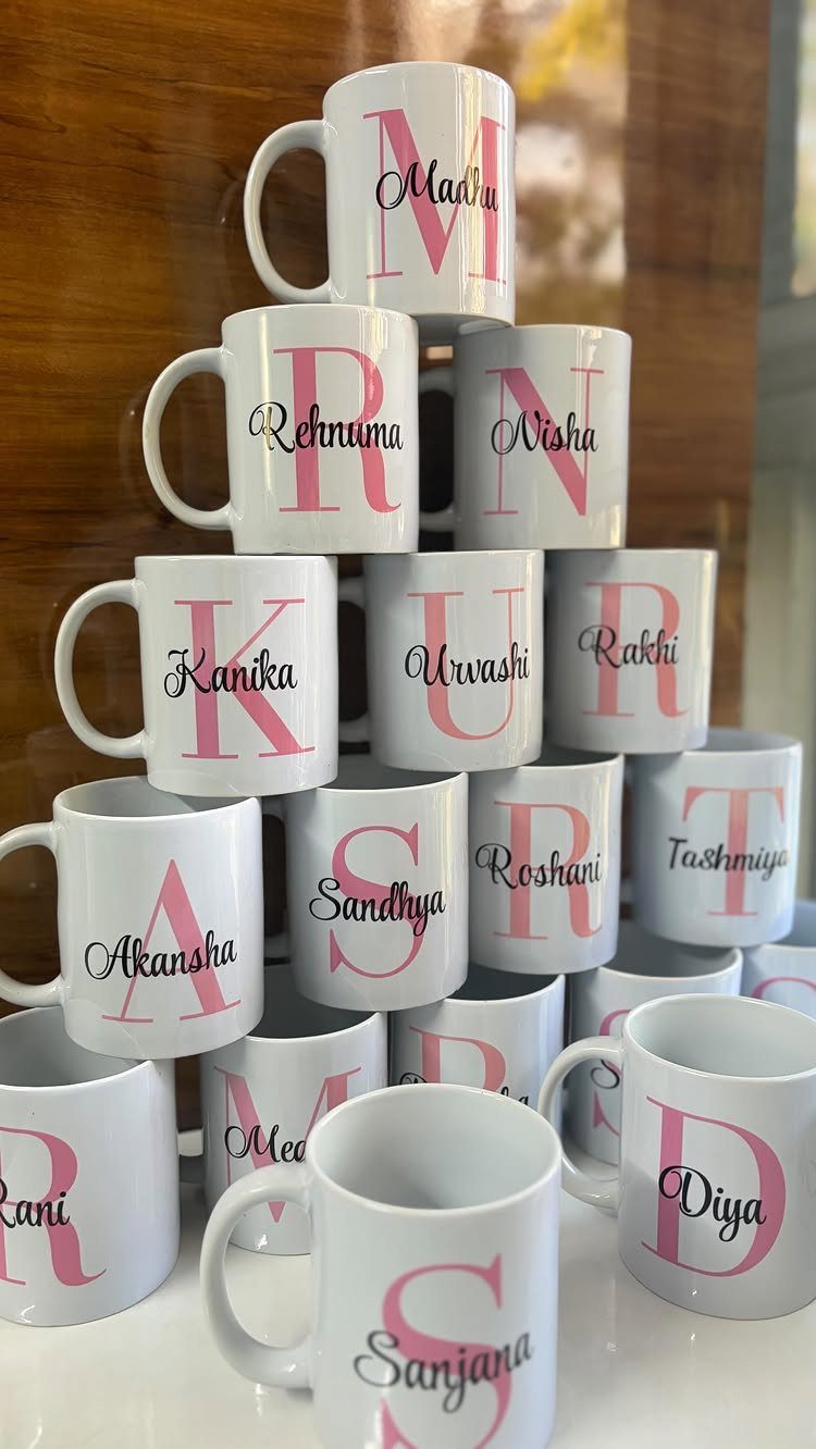 Stacked white ceramic mugs with pink initials and personalized names in black script.