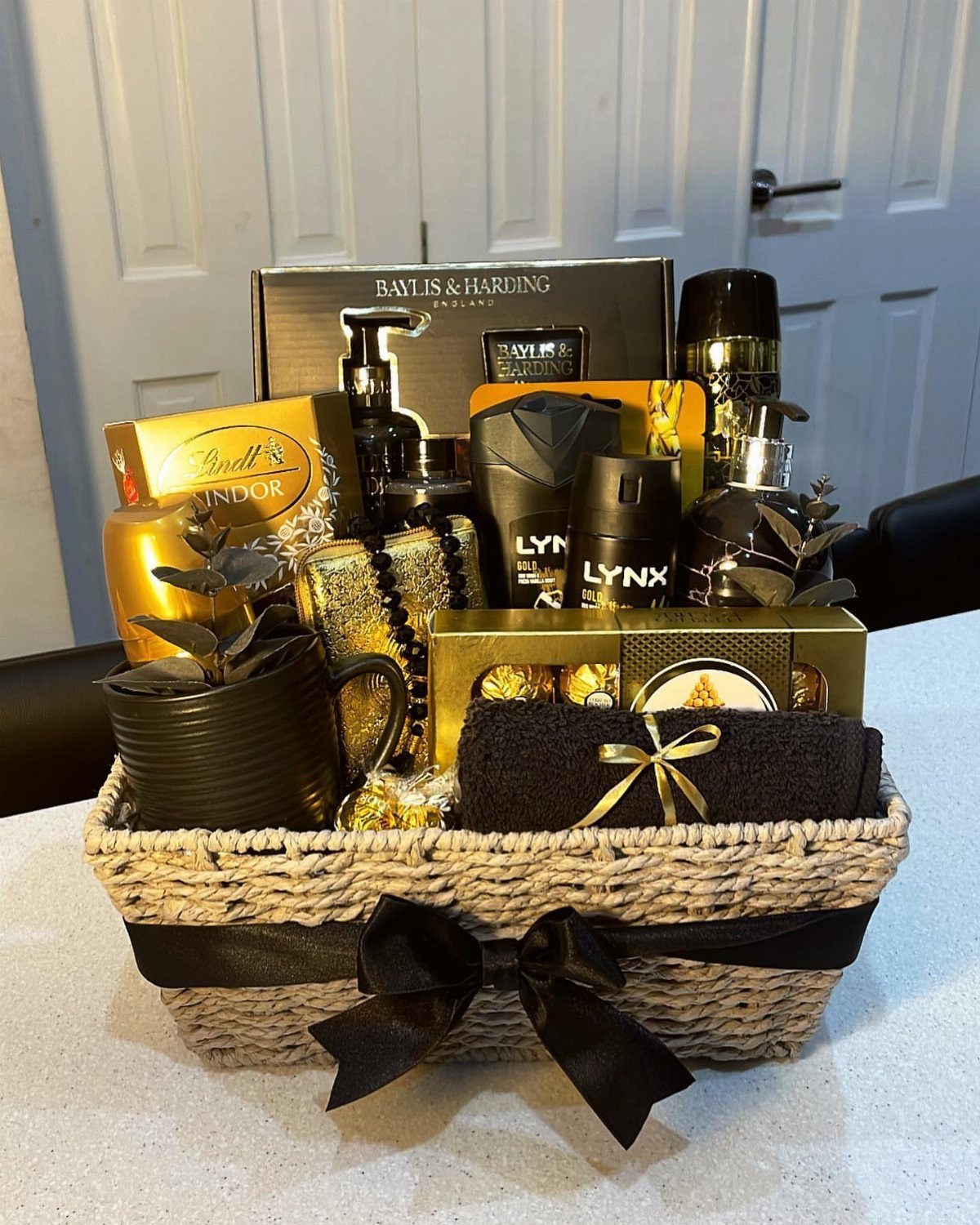 Woven gift basket filled with black and gold toiletries, chocolates, and a towel.
