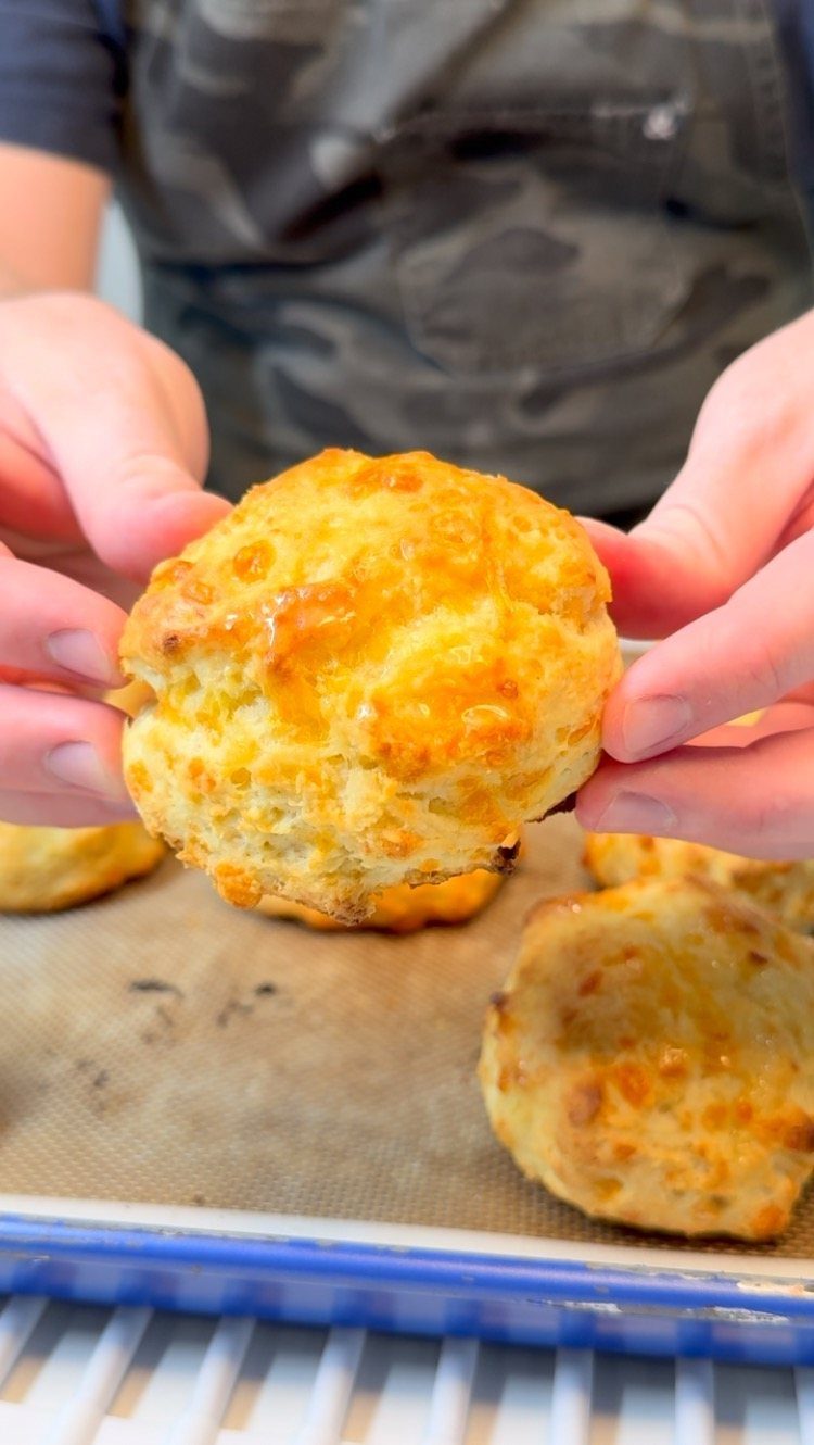 Golden-brown cheddar biscuit held by hands over a baking sheet of more biscuits.