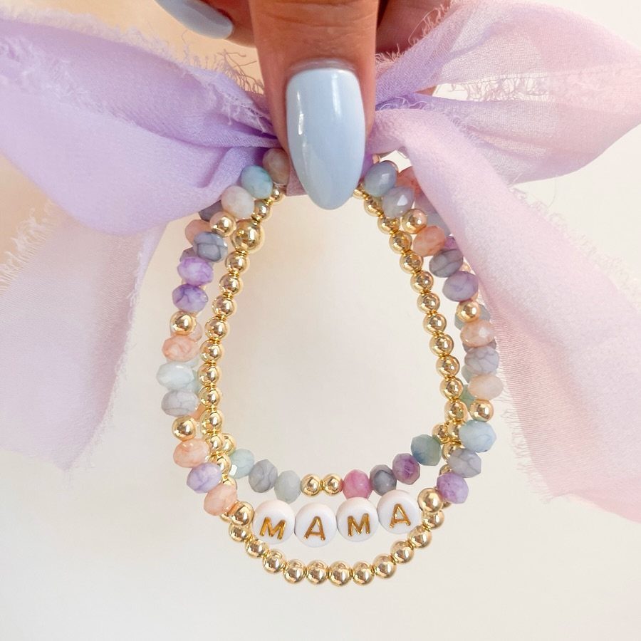 Two beaded bracelets, one pastel and one gold with MAMA letters, held by hand.