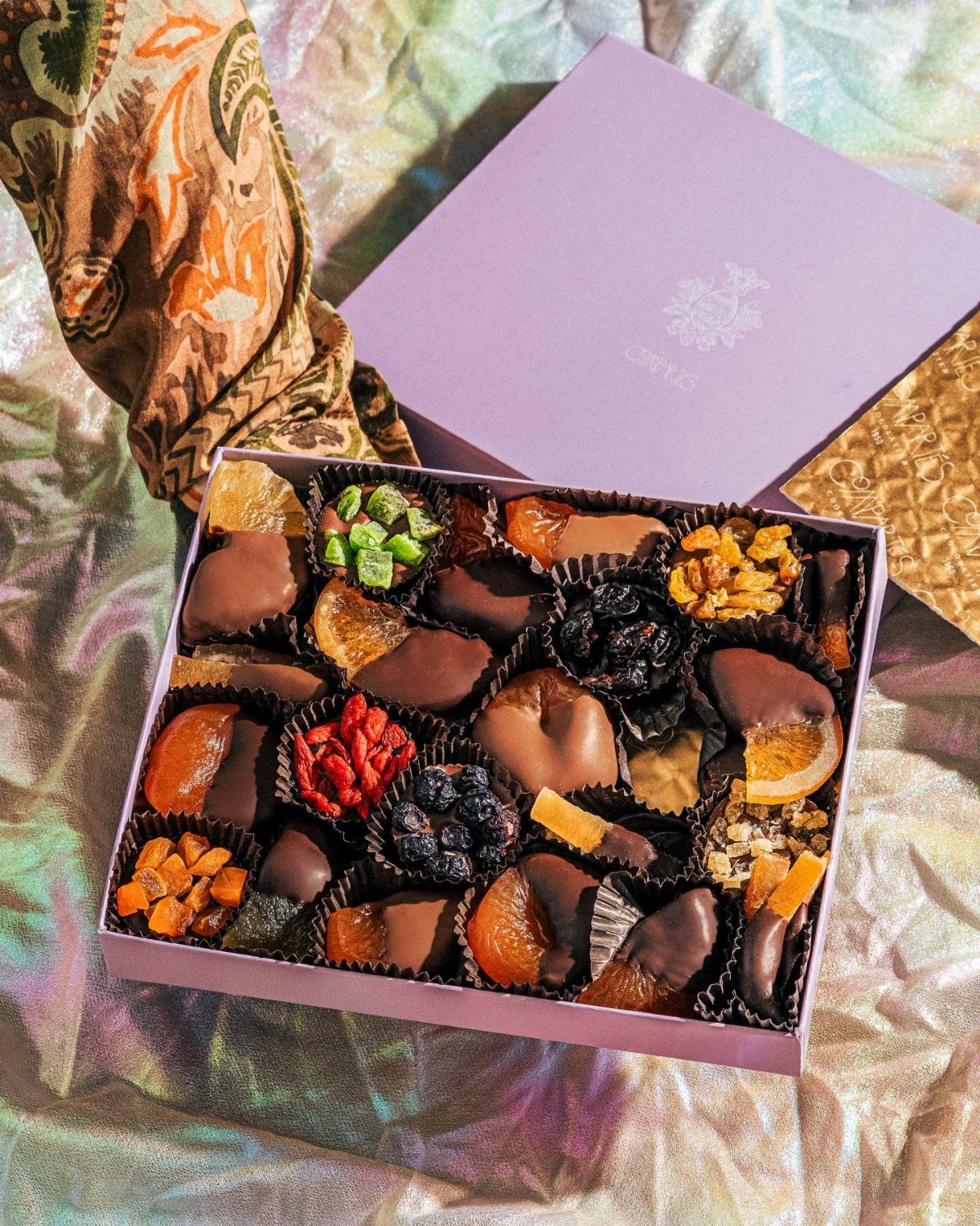 Assorted chocolate-dipped dried fruits and nuts in a purple gift box on iridescent fabric